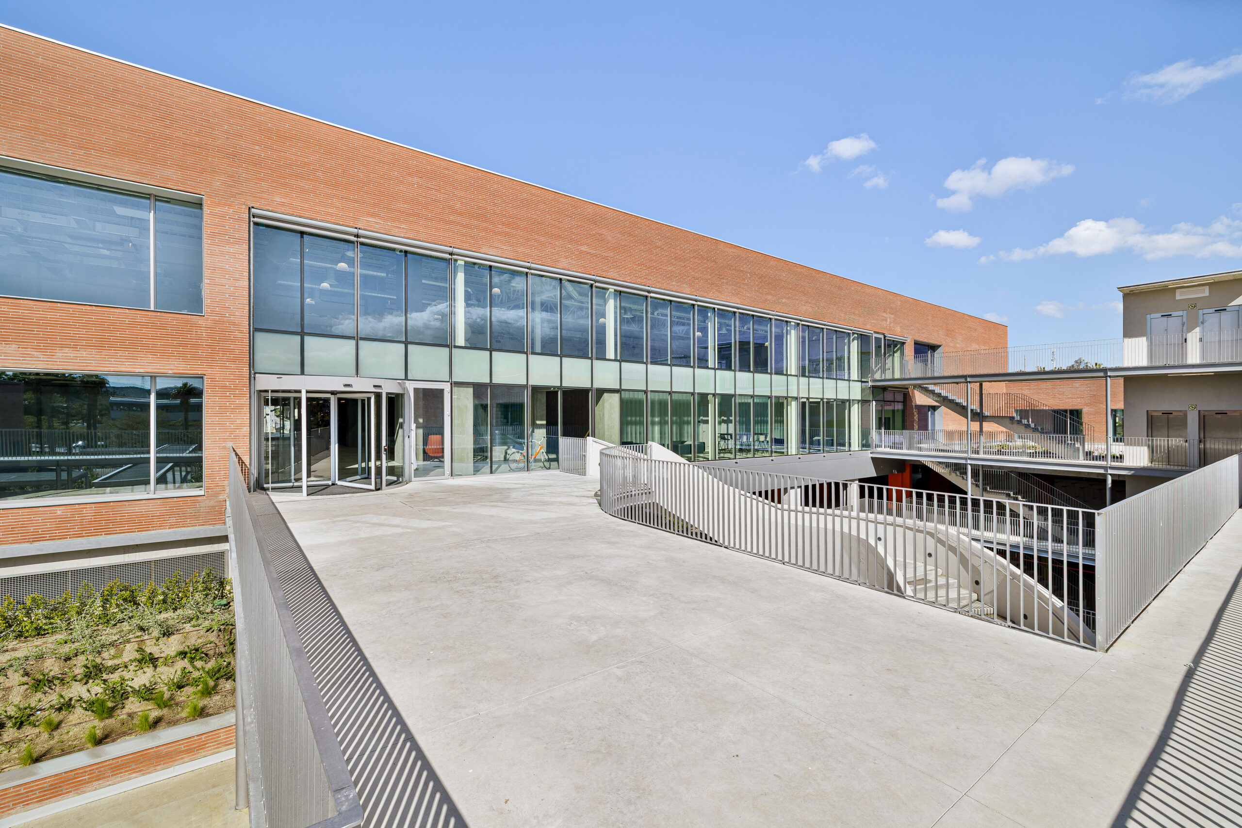 Glass-fronted entrance of Kadans NODE I in Málaga with a curved terrace, metal railings, and a red brick facade under blue skies.