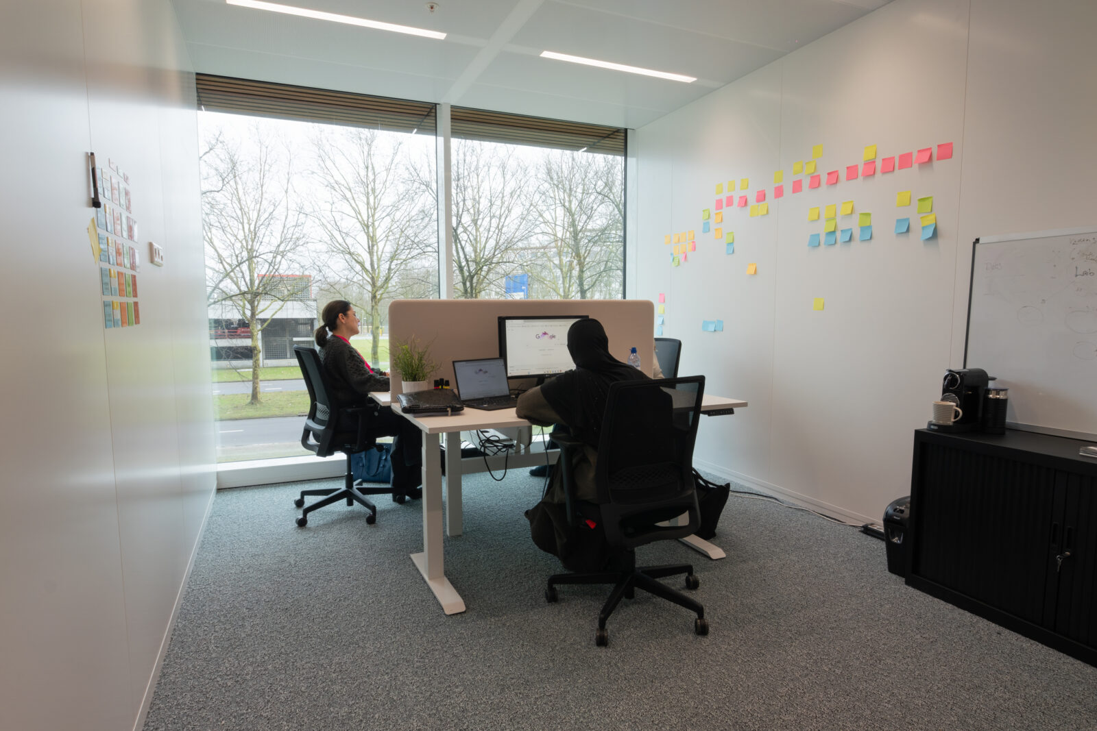 Two employees working at desks in Plus Ultra Utrecht with sticky notes on the wall - Kadans Sciene Partner